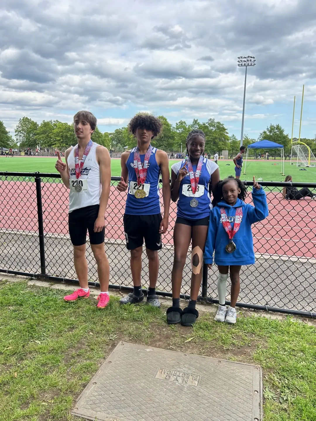 Four Metropolis Long Island Track Club athletes in club uniforms wearing medals at an outdoor track meet.